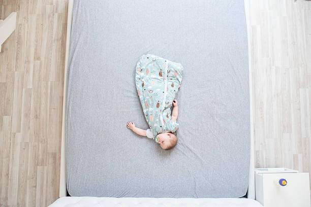 Adorable baby sleeping on a grey bed, secure in a patterned baby sleep sack.