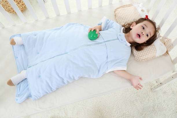 Toddler in a blue zippered baby sleep sack looking up from their crib.