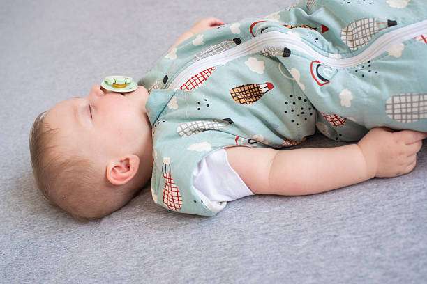 Baby sleeping soundly on its back wearing a blue patterned baby sleep sack with pacifier.