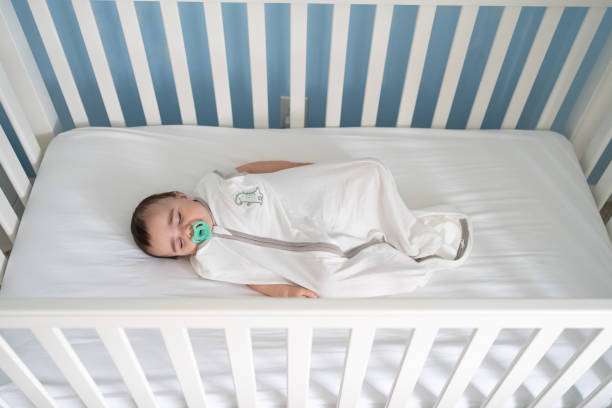 Peaceful baby sleeping on side in a crib, wearing a white baby sleep sack with a pacifier.